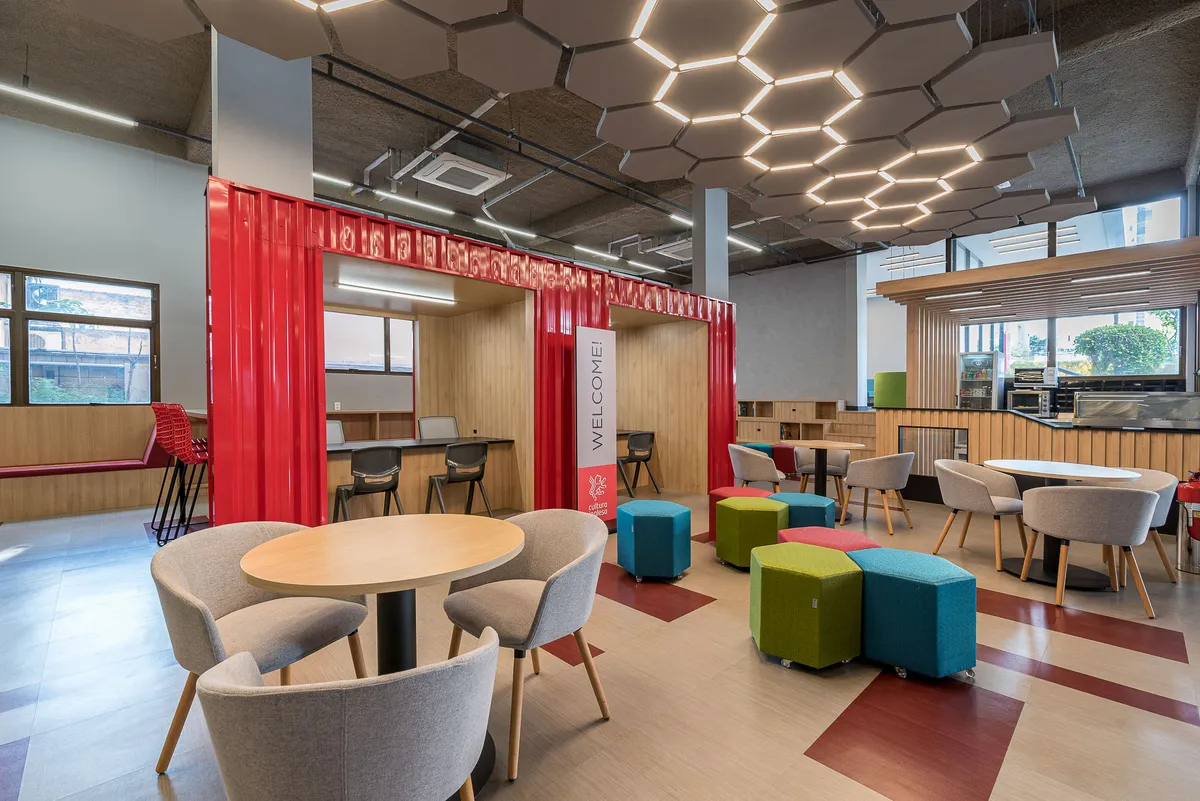 Área de convivência com luminárias hexagonais, poltronas cinza, mesa redonda de madeira e pufes coloridos.