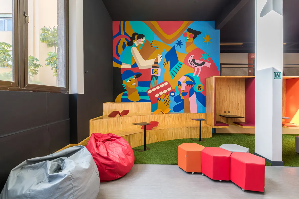 Espaço com mural ilustrado colorido, arquibancada com grama sintética, pufes vermelhos e laranja e puffs de tecido.
