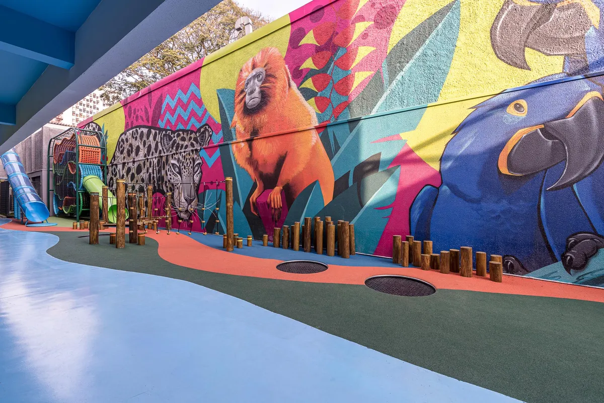 Playground com grande mural da fauna brasileira (onça-pintada, mico-leão-dourado e arara-azul), troncos de madeira e piso emborrachado colorido.