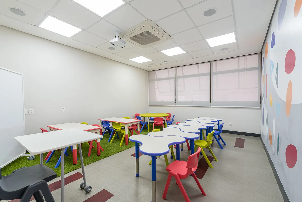 Sala da educação infantil com mesas modulares, cadeiras coloridas, tapete de grama sintética e parede com bolinhas coloridas.
