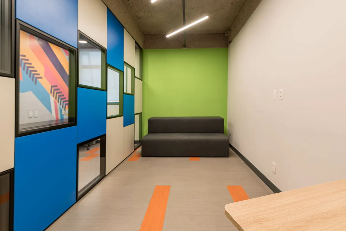Sala de estar com sofá preto, parede verde, painéis de vidro coloridos e piso com faixas laranja.