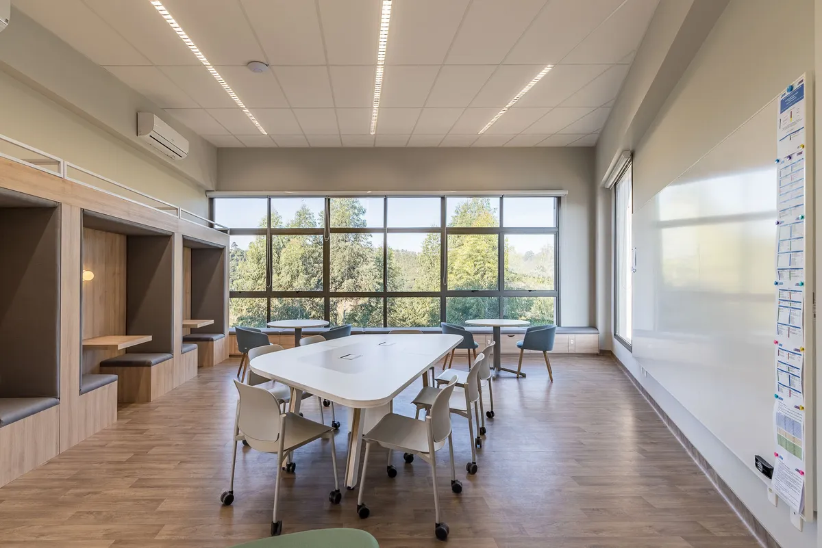 Sala de aula com mesa de trabalho coletiva branca, cadeiras com rodízios, cabines de estudo em madeira e janelas com vista para árvores.