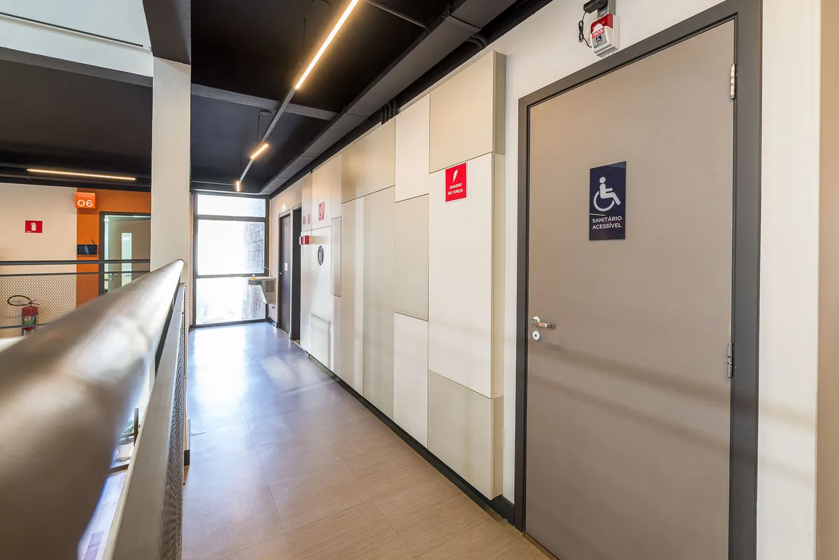 Corredor com painéis claros nas paredes, porta de banheiro acessível e guarda-corpo.