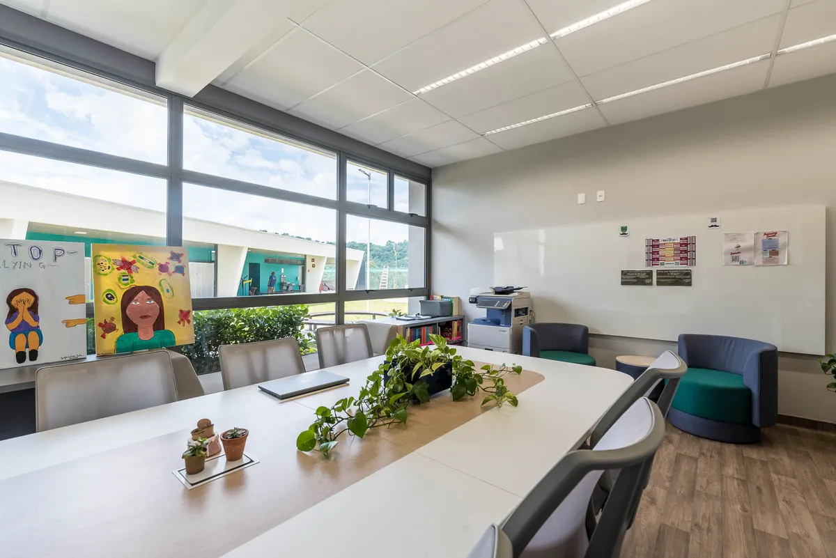 Sala dos professores com mesa coletiva branca de tampo de madeira, poltronas verde e azul e janelas amplas.