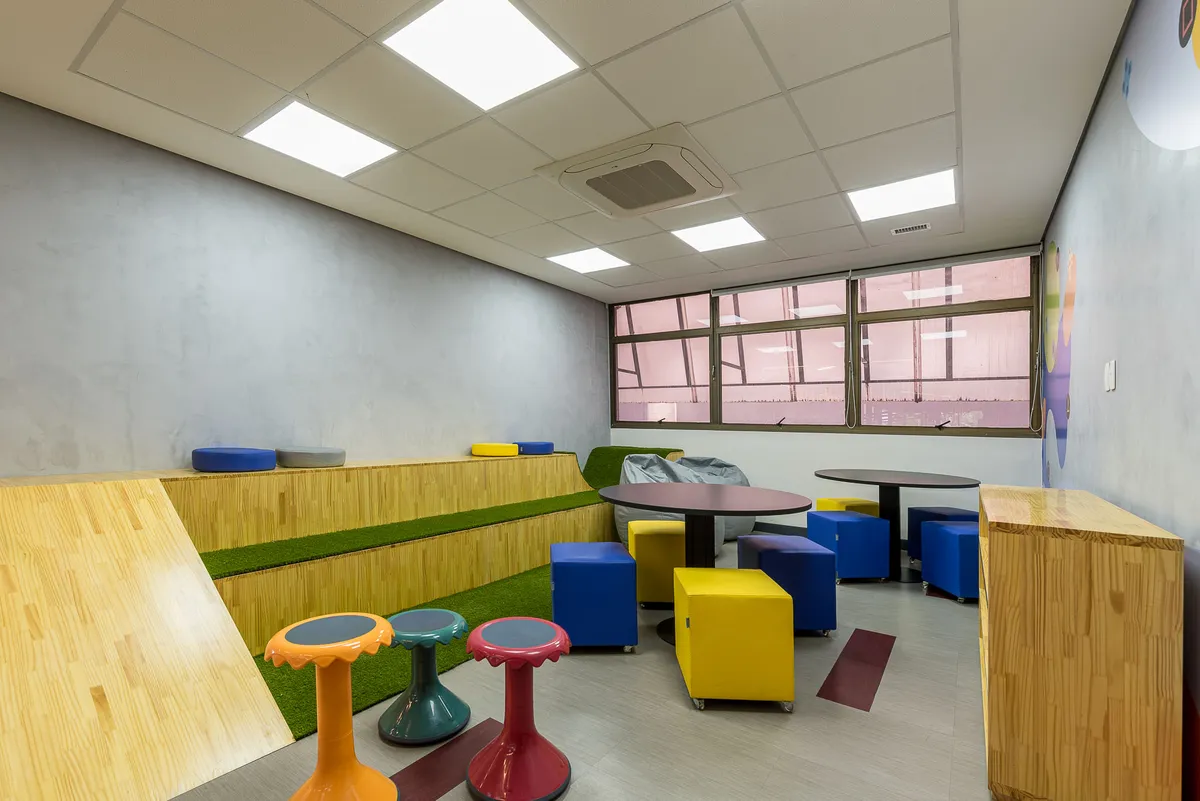 Espaço de convivência com arquibancada de madeira e grama sintética, pufes coloridos e banquetas.