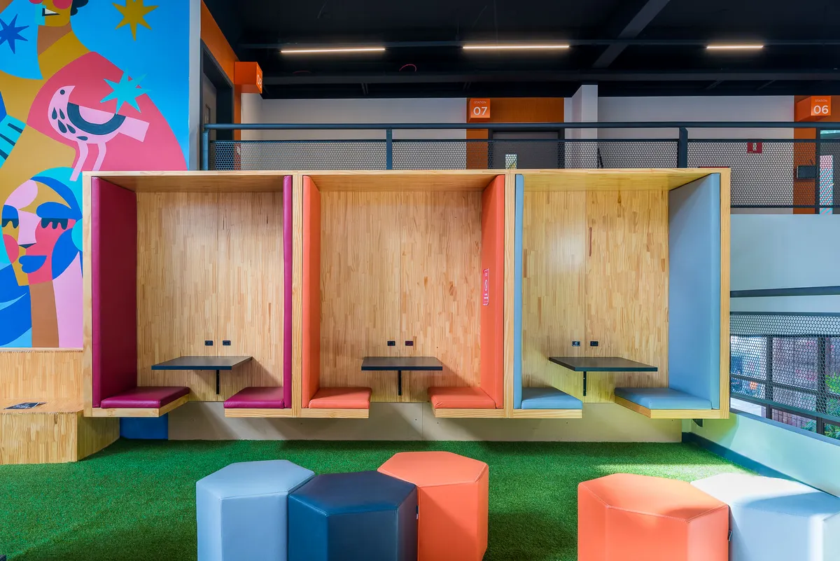 Cabines de estudo em madeira com bancos coloridos, grama sintética, pufes e mural ilustrado.
