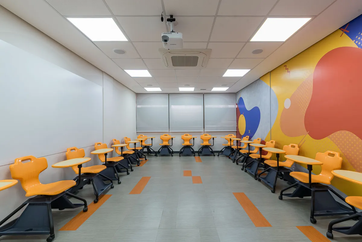 Sala de aula com cadeiras laranja com prancheta dispostas em U e mural colorido na parede.