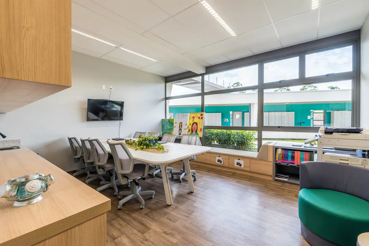 Sala dos professores com mesa coletiva, cadeiras de escritório, bancada de madeira e poltrona verde.