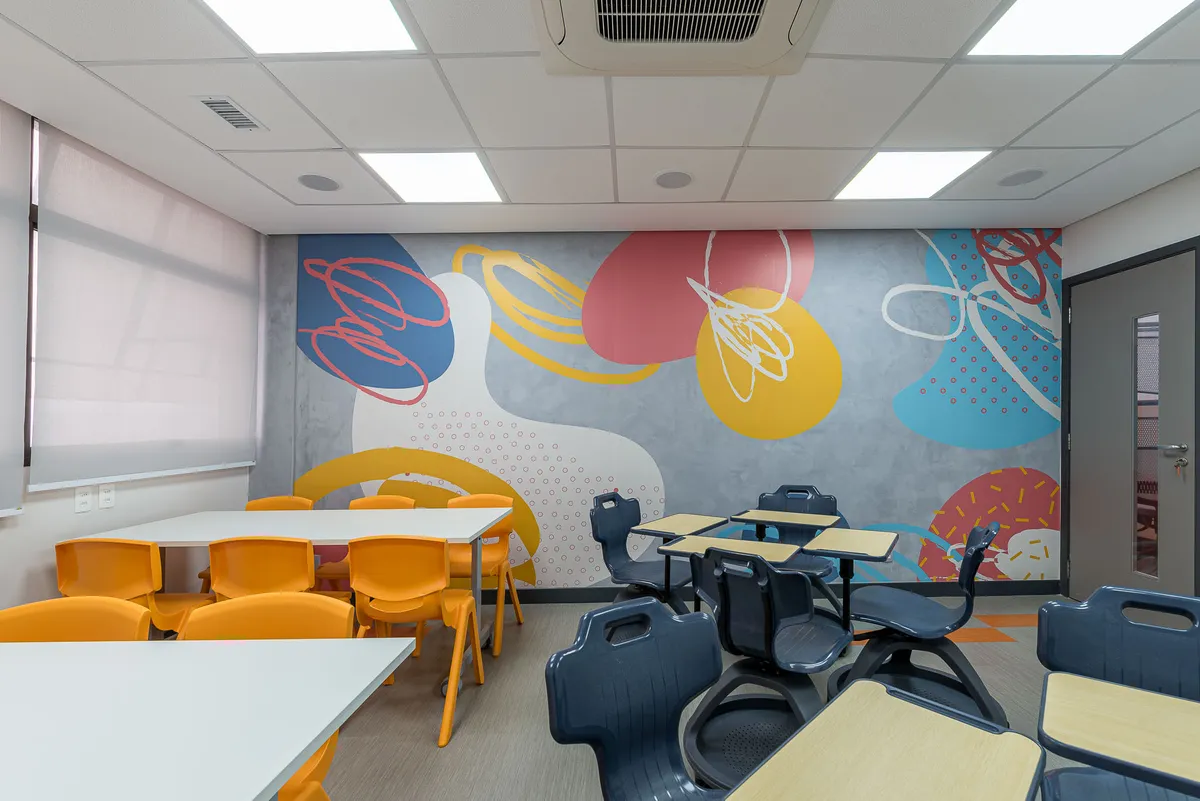 Sala de aula com cadeiras laranja e azul-marinho, mesas brancas e mural de formas coloridas.