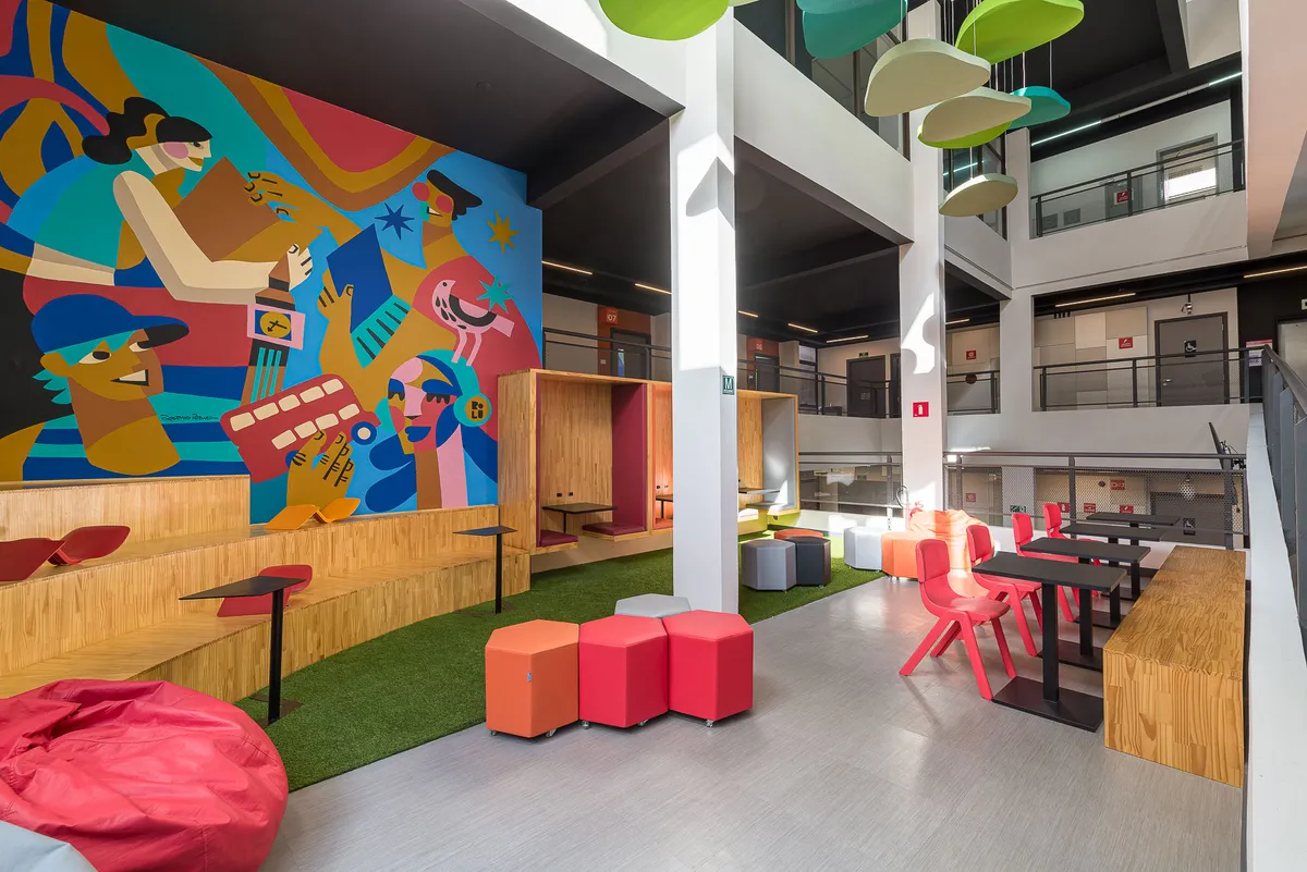 Área de convivência com grande mural ilustrado colorido, cabines de madeira, grama sintética e pufes.