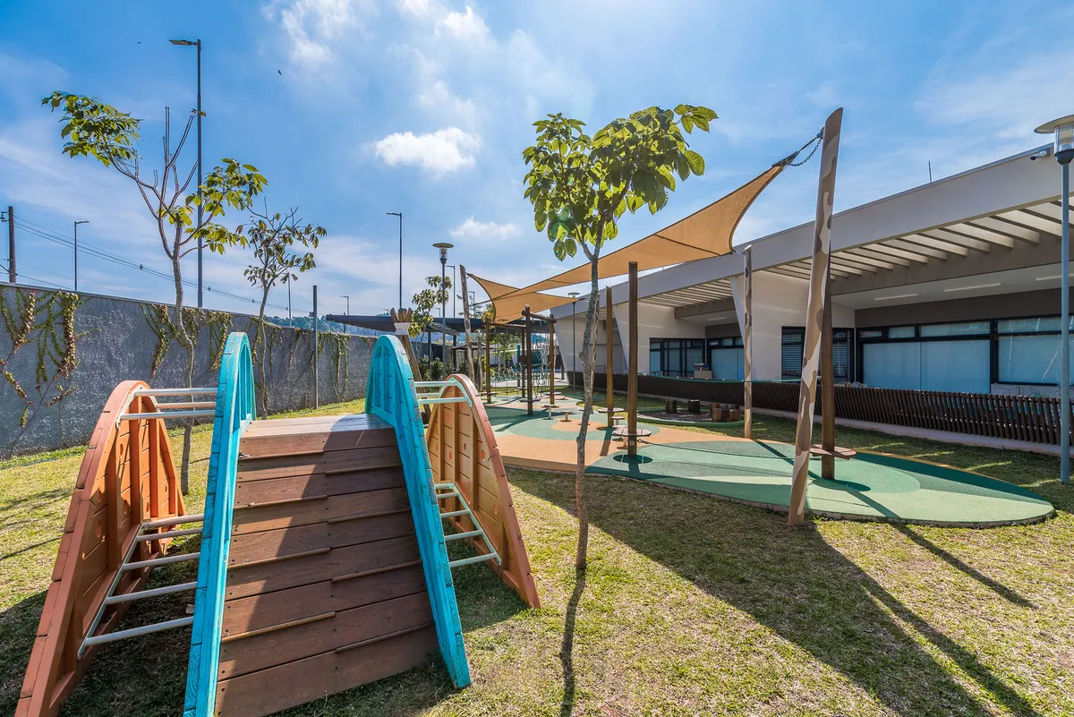 Playground externo com estrutura de escalada de madeira em arco, gramado e toldos tensionados.