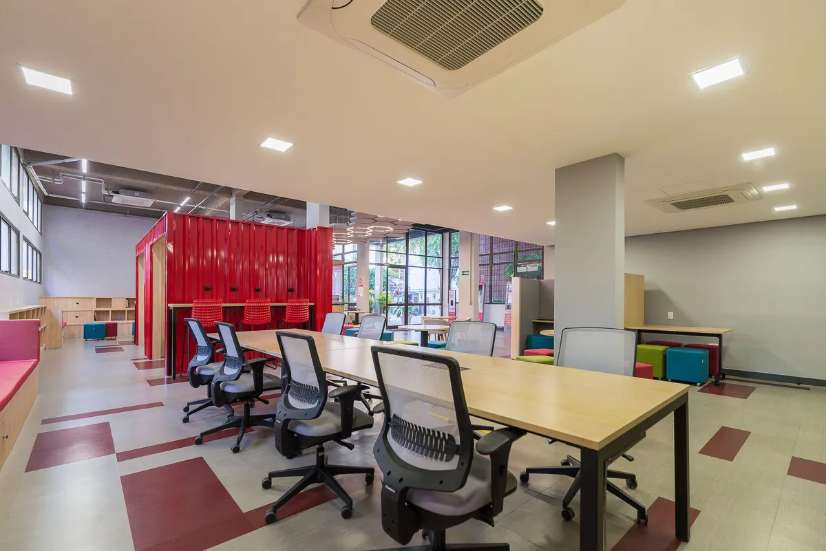 Sala de estudos com mesa longa de madeira, cadeiras de escritório, piso com faixas vermelhas e contêiner vermelho ao fundo.