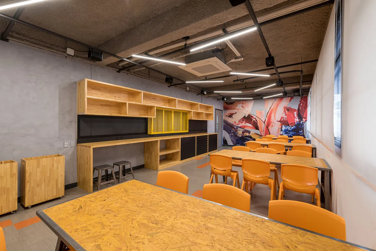 Laboratório maker com teto de concreto aparente, mesas de madeira, cadeiras laranja, marcenaria com nichos e mural grafitado.