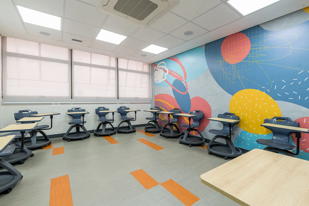 Sala de aula com cadeiras de rodízio azul-marinho com prancheta, mural de grafismos coloridos e piso com faixas laranja.