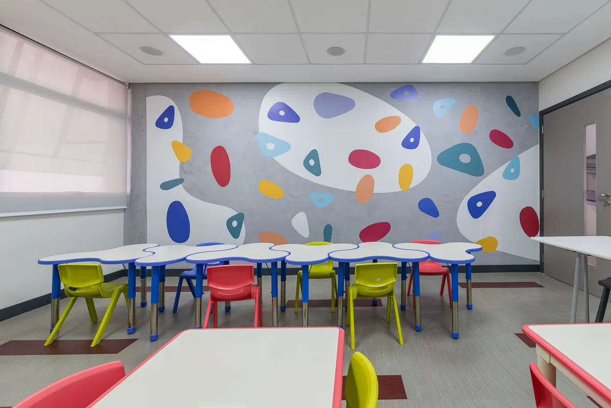 Sala de aula com mesas modulares brancas e azuis, cadeiras coloridas e parede com formas coloridas.