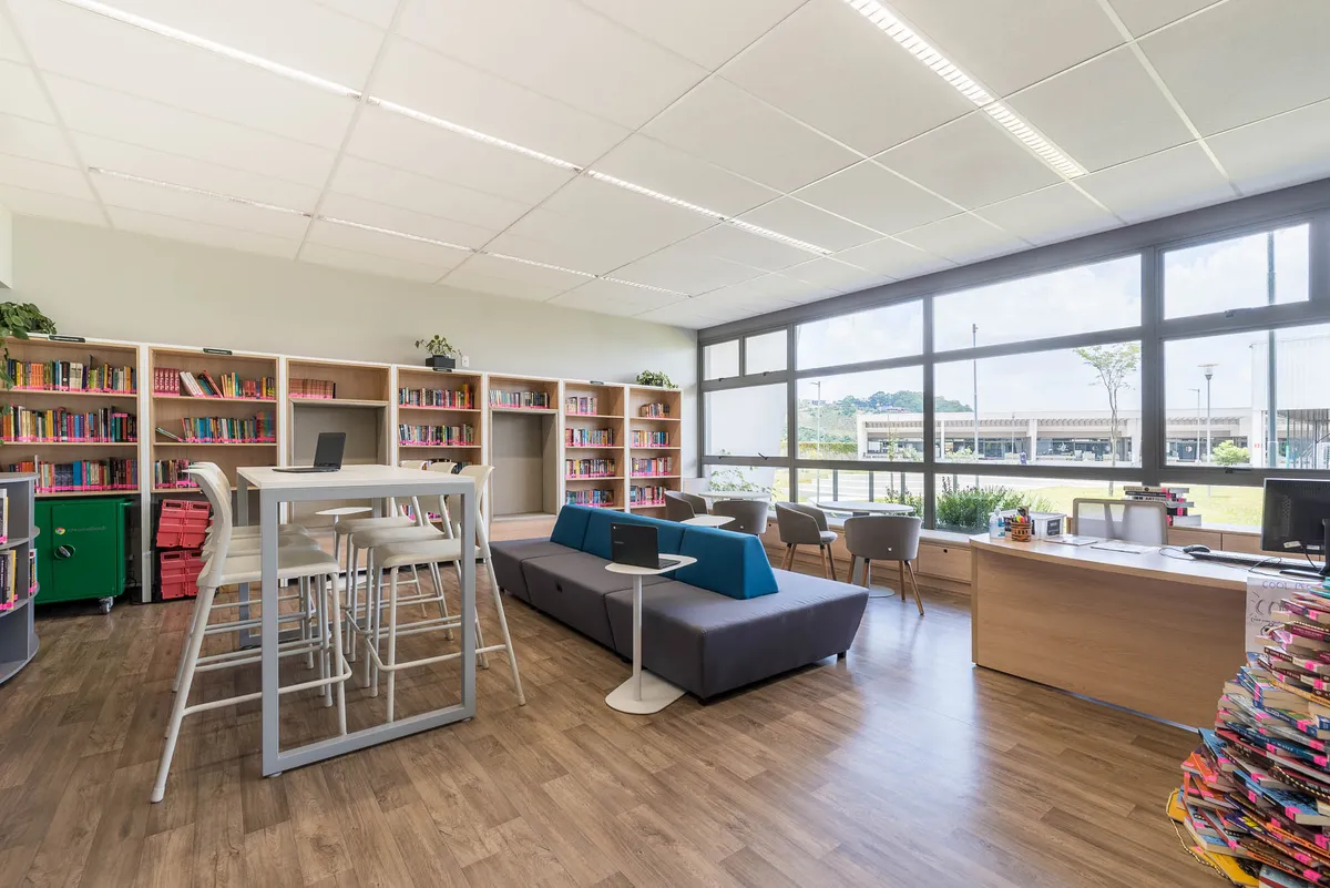 Espaço aberto com estantes de livros coloridas, mesas altas, sofá azul e mesa de atendimento.