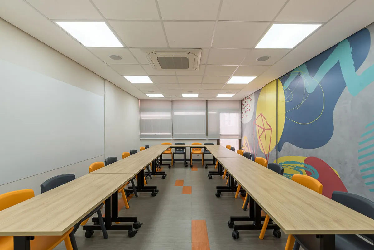 Sala de aula com mesas longas de madeira em fileiras, cadeiras laranja e pretas e mural colorido.