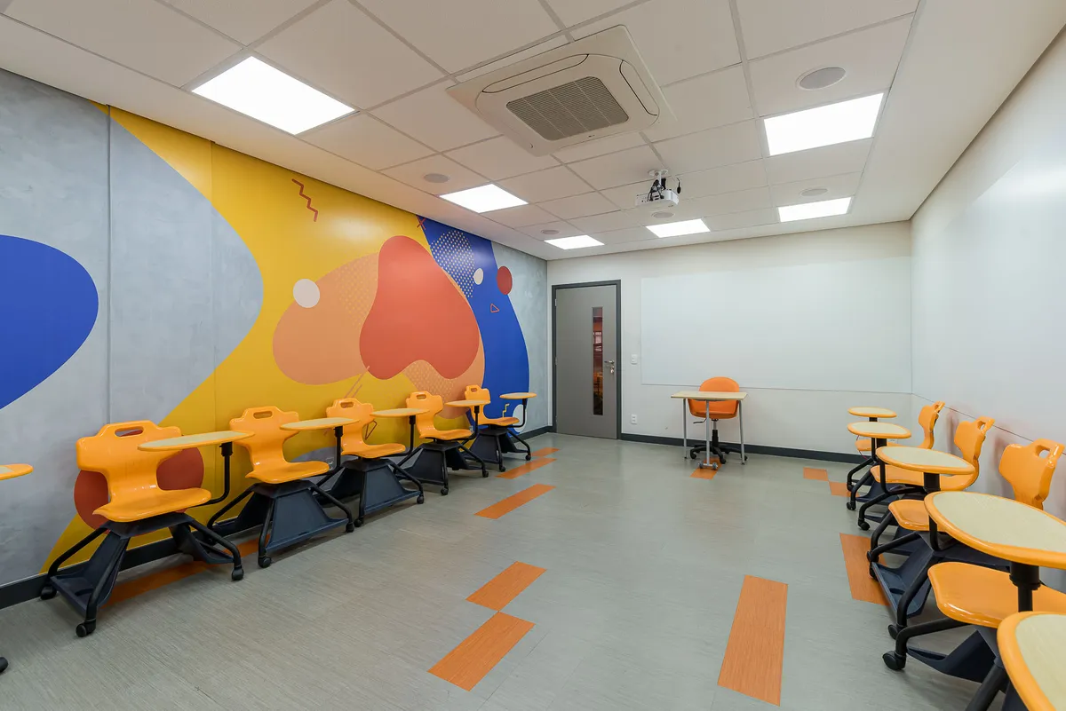 Sala de aula com cadeiras laranja com prancheta junto às paredes e grande mural de formas coloridas.