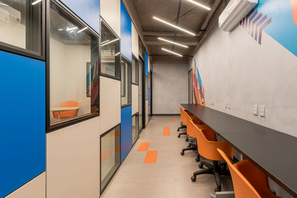Corredor com bancada de estudo, cadeiras laranja, salas envidraçadas com painéis azuis e parede cinza com grafismos.
